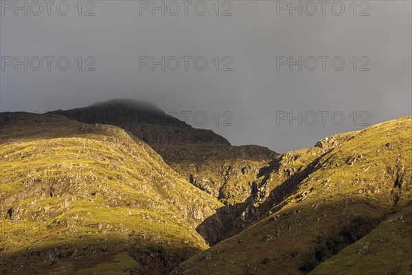 Rain cloud forming thick fog descending from steep mountainside bading in evening light at sunset in Glen Coe