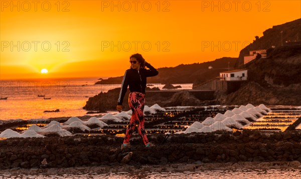 A young woman walking in the orange sunset in the salina and in the background the Fuencaliente Lighthouse on the route of the volcanoes south of the island of La Palma