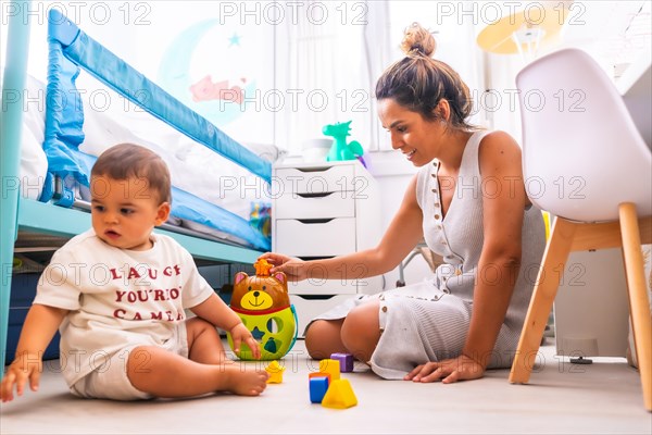 Young Caucasian mother playing with her in the room with toys. Baby less than a year learning the first lessons of her mother. Mother playing with her son playing sitting on the floor