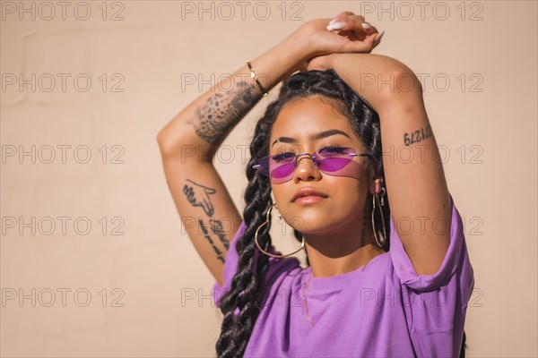 Urban session. Young dark-skinned woman with long braids wearing purple glasses on a plain cream-colored background