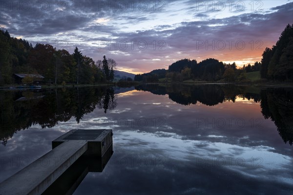 The Hengelesweiher pond in the Hengelesweiher nature reserve at sunset in autumn. A footbridge in the foreground. The lake is surrounded by forest. The sky with coloured clouds is reflected in the water. Isny im Allgaeu