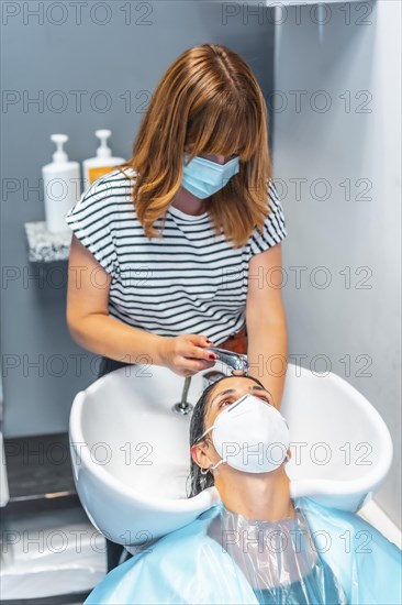 Hairdresser with face mask washing hair in the hot sink to the client. Safety measures for hairdressers in the Covid-19 pandemic. New normal