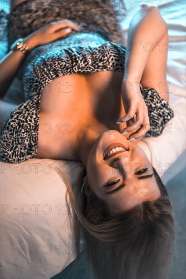 Lifestyle with a young blonde Caucasian woman in a short dress with a large black and white neckline lying on the bed on her back. Looking smiling