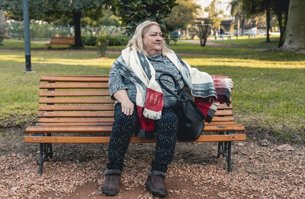 Senior serious woman waits for someone sitting on a bench at a public park