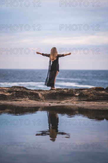 Summer lifestyle with a young brunette Caucasian woman in a long black transparent dress on some rocks near the sea on a summer afternoon. Raising arms and looking out to sea