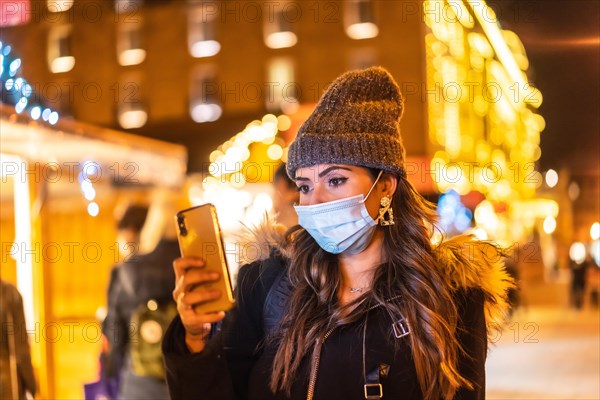 Christmas lifestyle in a new normal. Young girl with face mask visiting the christmas market looking at the mobile phone in the coronavirus pandemic