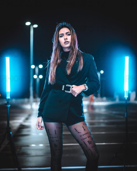 Caucasian brunette in pink wool jacket in an empty car park illuminated with blue light. Night urban session in the city