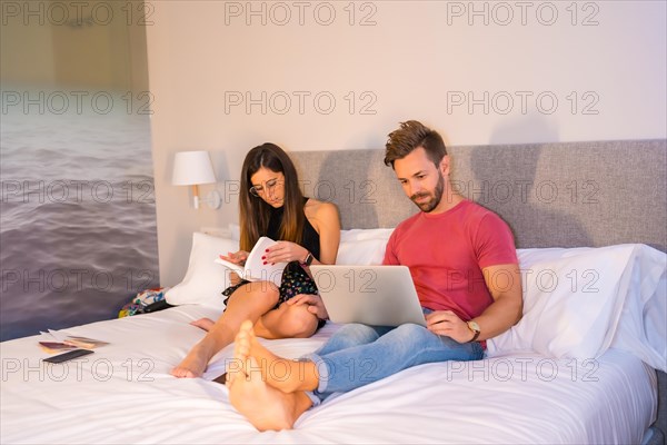 A Caucasian couple lounging on the hotel bed and looking at a computer and a book. Summer lifestyle