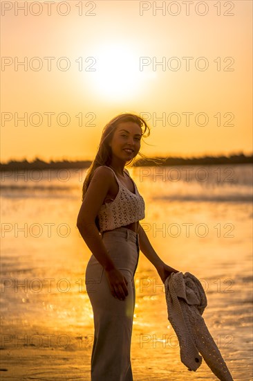 Summer lifestyle. A young blonde Caucasian woman in a white short wool sweater on a beach sunset. With eyes closed and enjoying the sea wind