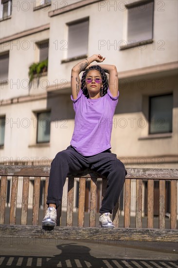 Young dark-skinned woman with long braids in purple glasses sitting on a bench in the park