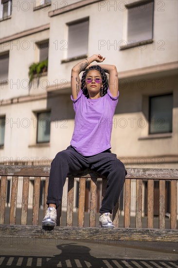 Young dark-skinned woman with long braids in purple glasses sitting on a bench in the park