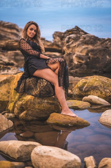 Summer lifestyle with a young brunette Caucasian woman in a long black transparent dress on some rocks near the sea on a summer afternoon. Sitting on a rock smiling and reflected in the water