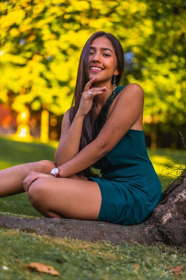 A young pretty brunette Latina with long straight hair leaning against a tree in a green dress. Portrait sitting on the grass next to a tree in the park