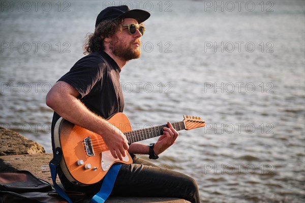 Hipster street musician in black playing electric guitar in the street sitting on pier embankment on sunset