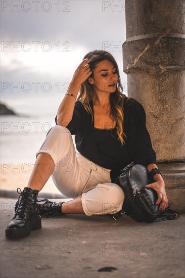 Lifestyle in the city with a blonde girl in white pants and a leather jacket near the beach. Sitting on the shallows of a beach at sunset