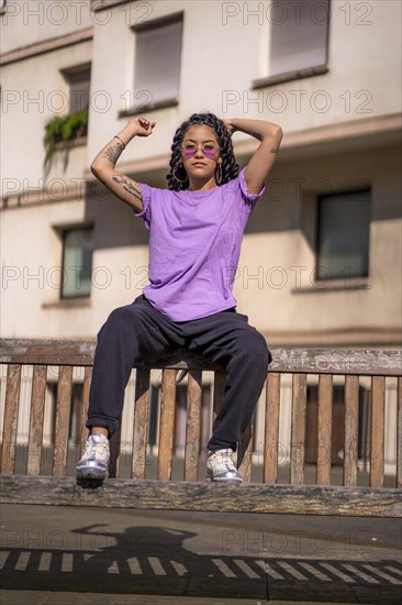 Young dark-skinned woman with long braids in purple glasses sitting on a bench in the park
