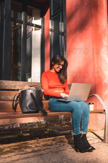 Lifestyle. Young caucasian brunette in a red shirt sitting on a bench in the park with a take away coffee. Working with the computer