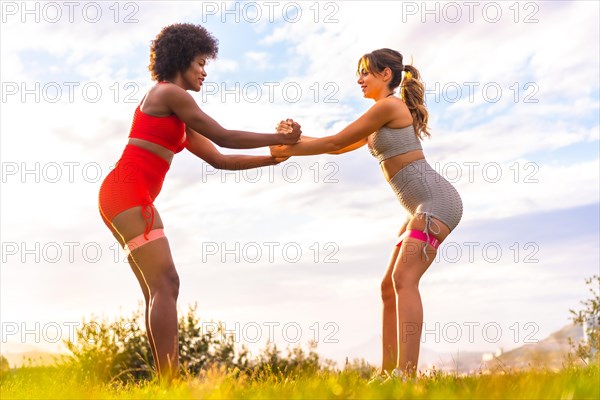 Caucasian blonde girl and dark-skinned girl with afro hair doing squat exercises in a park with the city in the background. Healthy life