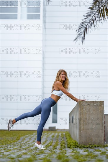 Fitness and yoga session with a young blonde Caucasian instructor dressed in a casual outfit with blue Maya and a white T-shirt. Exercising with a white wall in the background