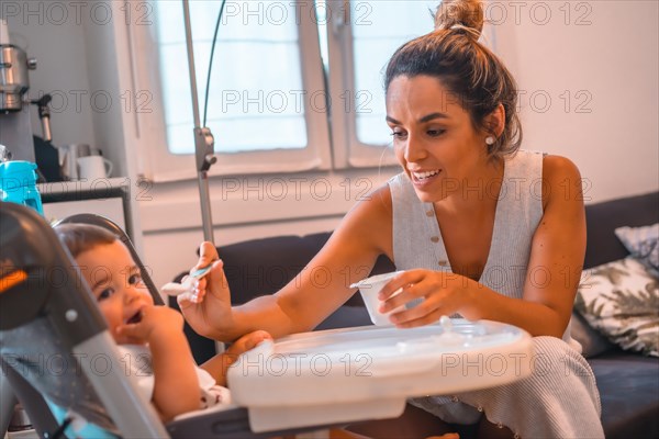 Young Caucasian mother feeding her son a yogurt while sitting in the highchair. Teleworking and caring for your child