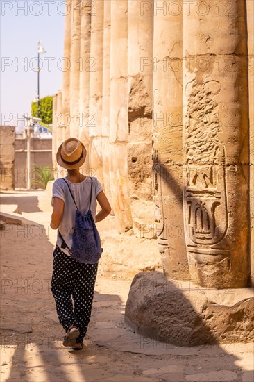 A young tourist in a white t-shirt and hat visiting the temple and looking at the ancient egyptian drawings on the columns of the Luxor Temple