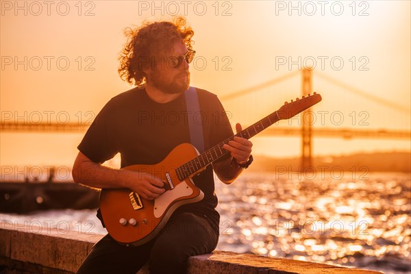 Hipster street musician in black playing electric guitar in the street on sunset on embankment with 25th of April bridge in background. Lisbon