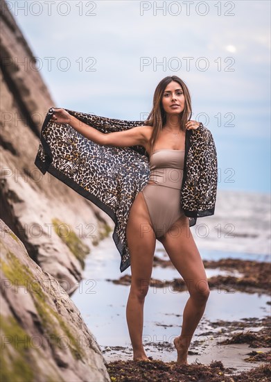 A dark-haired caucasian woman in a brown swimsuit and sarong on a natural background next to rocks and sea. In the summer sunset. Posing with the beautiful sarong next to the water