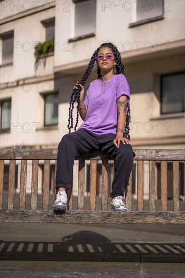 Young dark-skinned woman with long braids in purple glasses sitting on a bench in the park