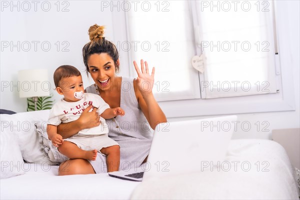 Young Caucasian mother with her son in the room on top of the bed. Drink less than a year in a video call with her family. Communication with grandparents in the confinement of the covid-19 pandemic