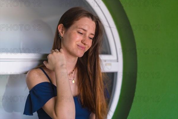 A young pretty redhead Caucasian girl smiling sitting in a blue dress next to a white sale of a green house