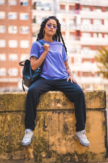 Dark-skinned young woman with long braids and purple glasses sitting on a bench in the city