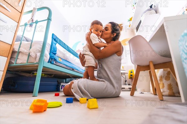 Young Caucasian mother playing with her in the room with toys. Baby less than a year learning the first lessons of her mother. Mother playing with her son and hugging him lovingly