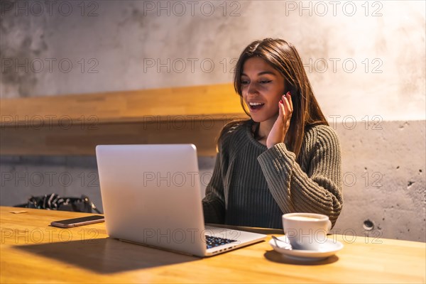 A young Caucasian brunette entrepreneur working from a coffee shop. Having a coffee and making a video call with the computer