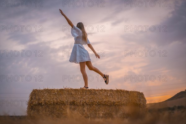 A young blonde Caucasian woman in a white dress in a field of dry straw atop a haystack. In a dry cultivated field in Navarra