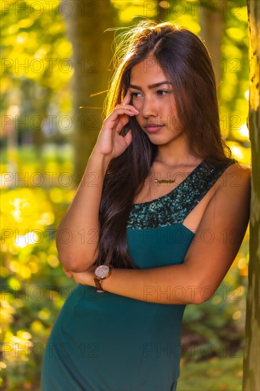 A young brunette Latina with long straight hair leaning against a tree in a green dress. Portrait with a sweet and sensual look