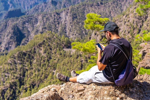 A young man resting after trekking at the top of La Cumbrecita sitting in the natural viewpoint and taking a photo with the mobile