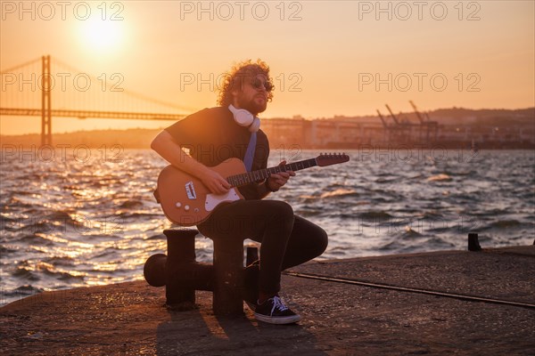 Hipster street musician in black playing electric guitar in the street on sunset on embankment with 25th of April bridge in background. Lisbon