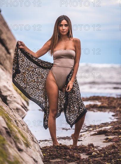 A dark-haired caucasian woman in a brown swimsuit and sarong on a natural background next to rocks and sea. In the summer sunset