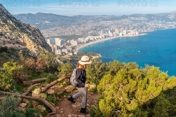 A young hiker wearing a hat on the descent path of the Penon de Ifach Natural Park with the city of Calpe in the background