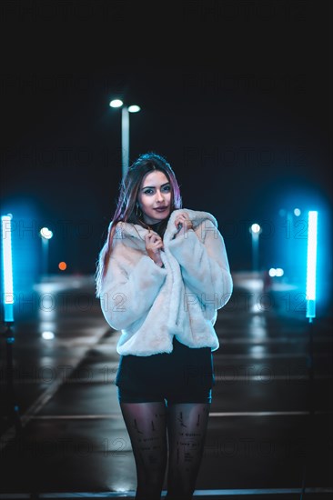 Young Caucasian brunette with pink wool jacket in an empty car park illuminated with blue light. Night urban winter session in the city