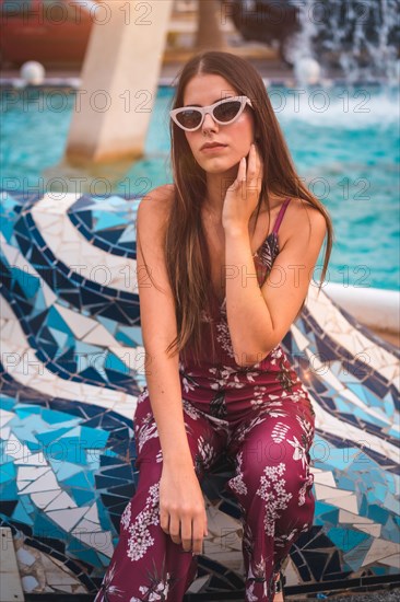 Portrait of a young brunette in a maroon floral dress enjoying the summer in the golden hour in a city fountain