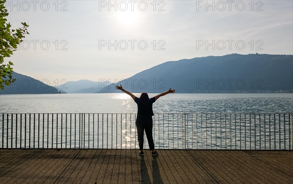 Woman with Shadow Standing with Arms Outstretched on the Waterfront with Railing to Lake Lugano with Mountain in a Sunny Summer Day in Bissone