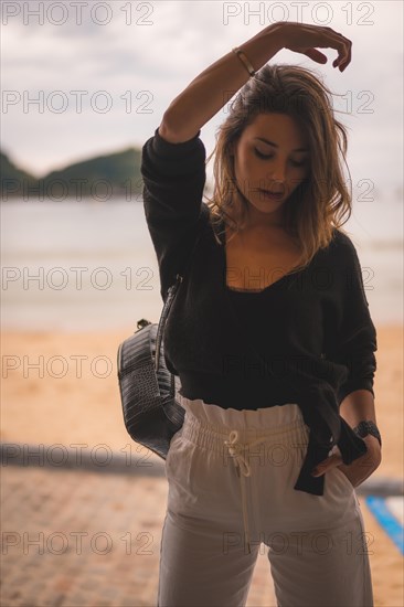 Lifestyle in the city with a blonde girl in white pants and a leather jacket near the beach. Very happy in some shallows on the beach in a sunset