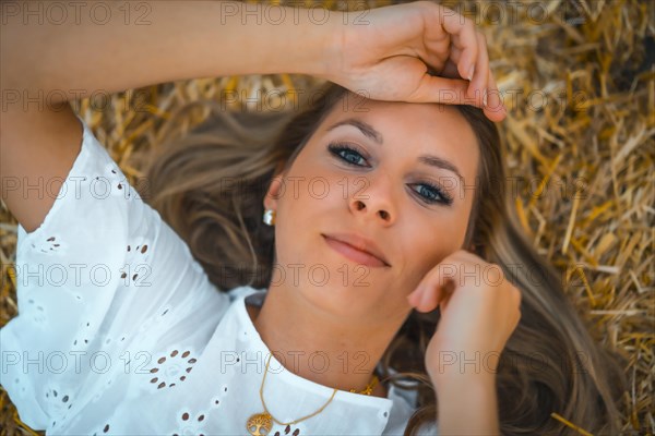 A young blonde Caucasian woman in a white dress in a field of dry straw atop a haystack