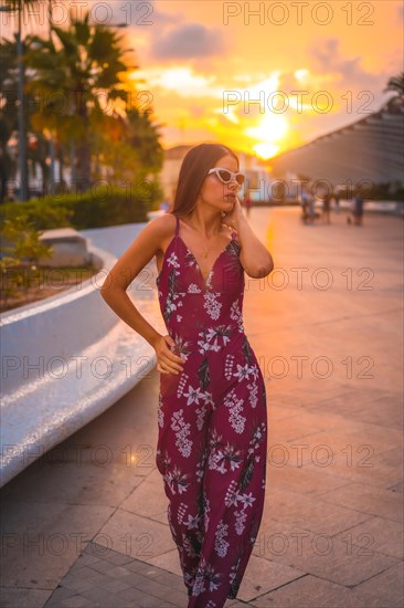 Sunset with a young brunette in a maroon floral dress and modern white sunglasses enjoying the summer in the golden hour