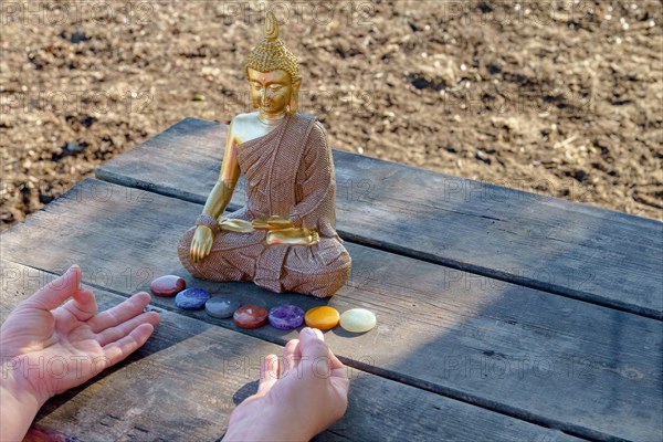 Woman's hands meditating in front of a Buddha statue with colorful chakra stones on a wooden table in a park in the sunshine