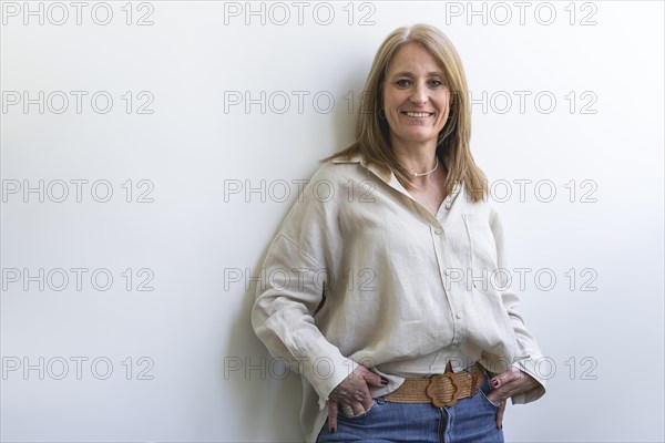 Beautiful caucasian mid aged woman looking at camera leaning on white wall