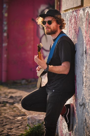 Hipster street musician in black playing electric guitar in the street on sunset leaning on a wall