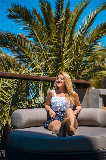 Summer lifestyle of a young blonde Caucasian woman in a white flowery top enjoying the summer on a terrace with palm trees. Sitting on a sofa on a terrace sunbathing and smiling