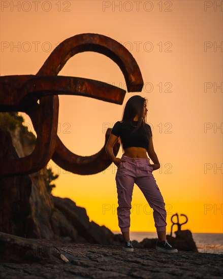 A young pretty brunette Latina with long straight hair in a short black T-shirt and pink pants. Orange sunset next to a famous sculpture of San Sebatian called Peine del Viento